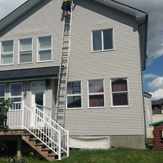 Roofer climbing an extension ladder to the second storey of a grey-sided house under a heavy cloud bank