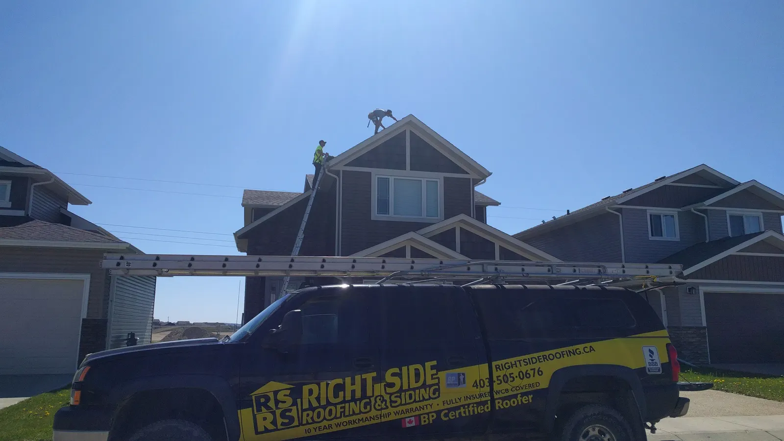 Truck at the curb with the ladder up against the eave and two roofers working on the ridge above