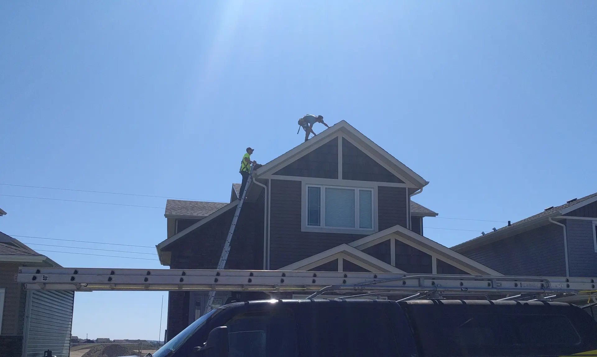 Truck at the curb with the ladder up against the eave and two roofers working on the ridge above